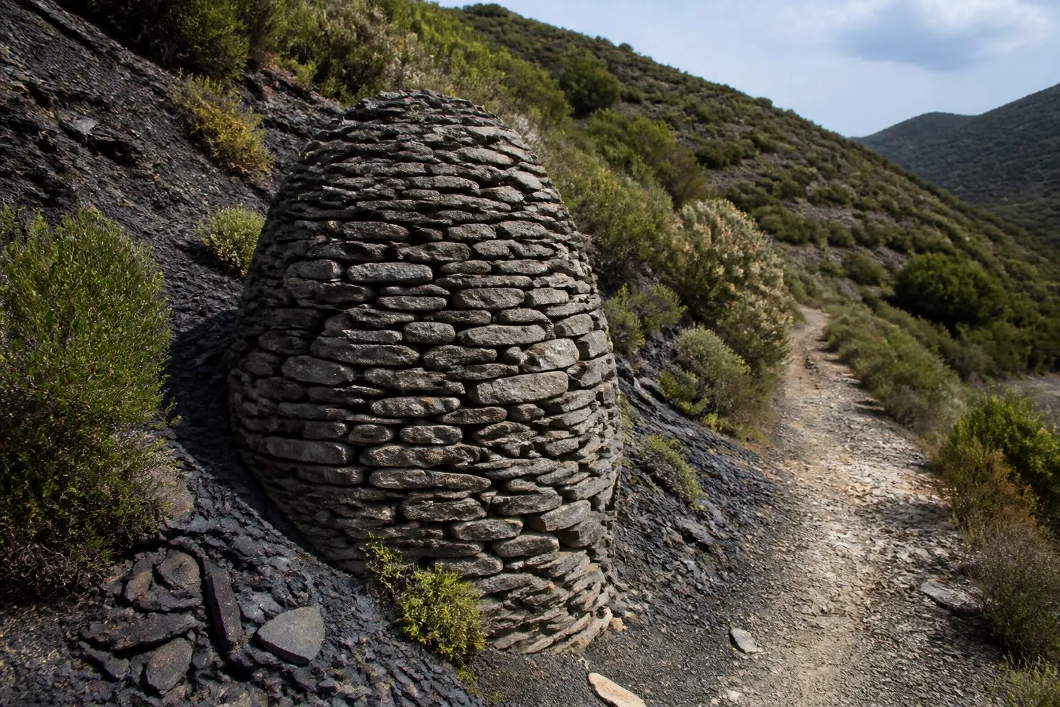 un cairn ovoïde de pierre sèche au bord d'un chemin de montagne, sans mortier apparent, entouré de marnes noires et de garrigue